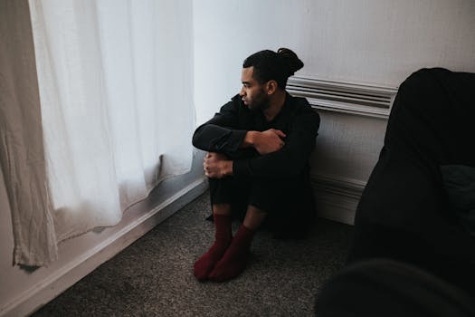 A contemplative man sits in a corner of a dimly lit bedroom, reflecting deep emotions.