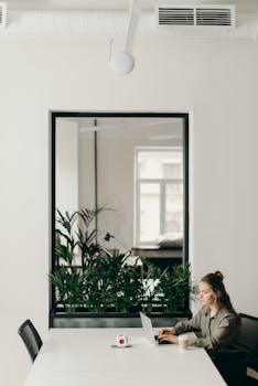 A young woman working on a laptop in a minimalistic, modern office setting with plants and a cup of coffee.