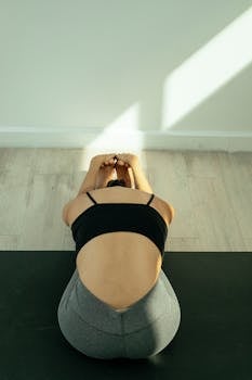 From above of unrecognizable fit female in activewear sitting on floor while stretching body in light apartment