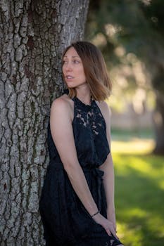 Portrait of a woman in a black dress leaning on a tree in a serene outdoor setting.