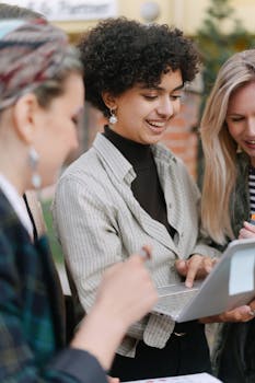 Three adult women collaborating on a laptop in an outdoor setting, smiling and engaged.